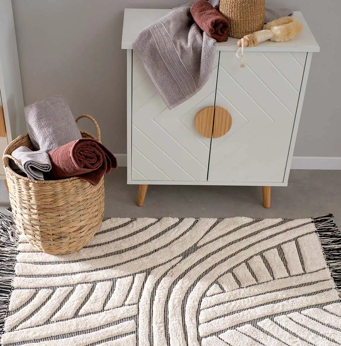 Tapis de salle de bain aux couleurs claires et neutres avec un motif géométrique, accompagné d’un panier en osier contenant des serviettes et d’un meuble blanc avec des poignées en bois et des serviettes pliées sur le dessus.