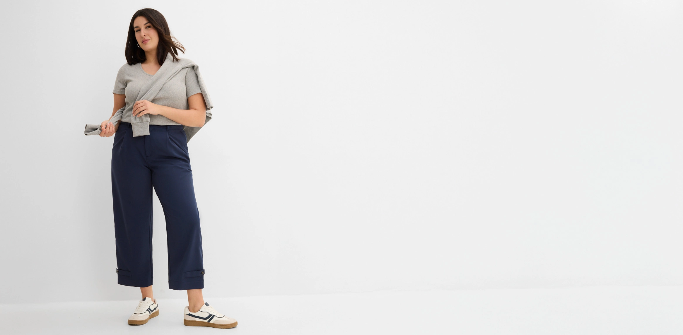 Look féminin avec un t-shirt gris, un pantalon bleu marine à panneau décoratif à la taille, un pull et des baskets blanches.