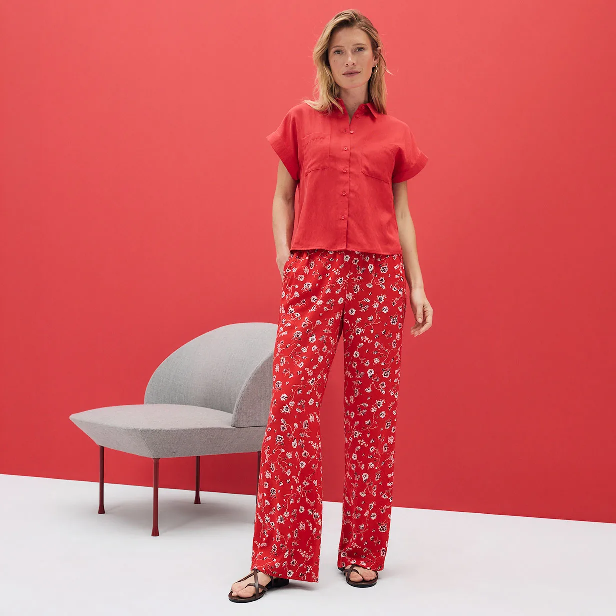 Chemisier rouge à manches courtes et pantalon fluide à motif floral. Une tenue estivale sur un fond rouge lumineux.