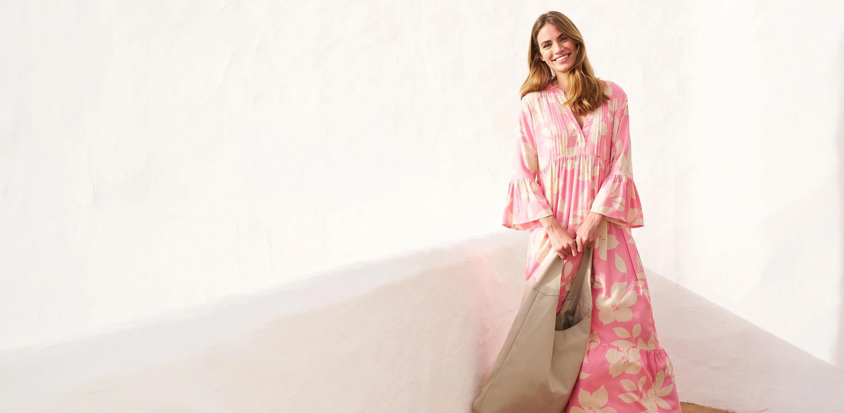 Une femme portant une robe fluide rose à fleurs, un grand sac beige et des baskets.