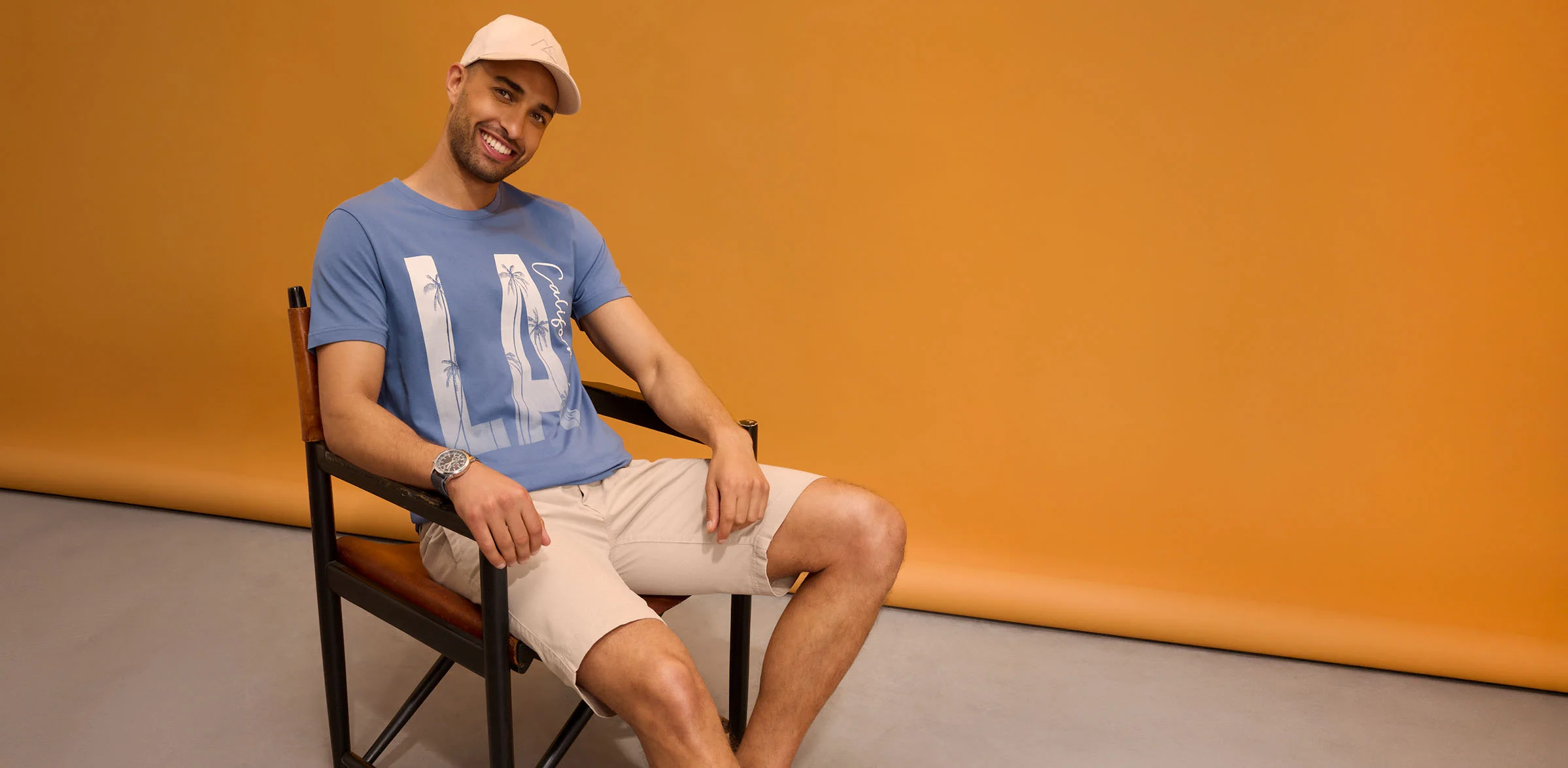 Look décontracté : casquette, T-shirt bleu à imprimé, short beige et montre.