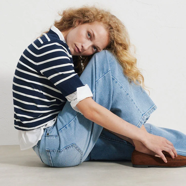 Pull rayé porté sur une chemise blanche, avec un jean large bleu et des mocassins marron.