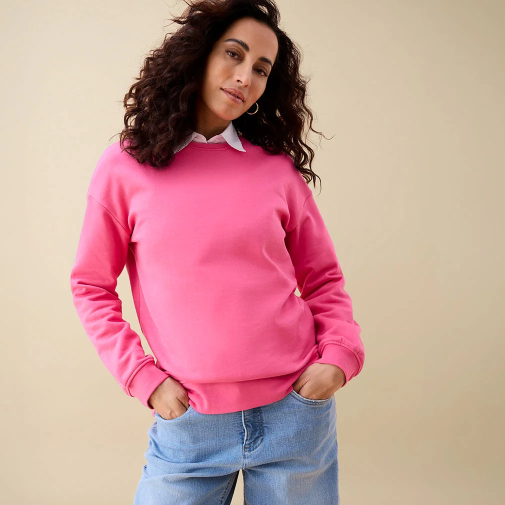 Sweat rose vif porté sur une chemise blanche, avec un jean bleu décontracté.