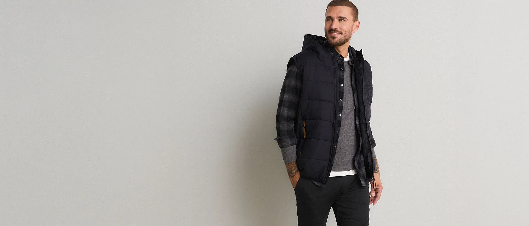 Veste matelassée foncée à capuche, chemise à carreaux, T-shirt gris et pantalon noir.