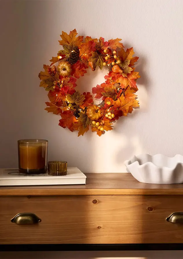 Couronne lumineuse automnale au mur, bougies et coupelle déco sur un meuble en bois.