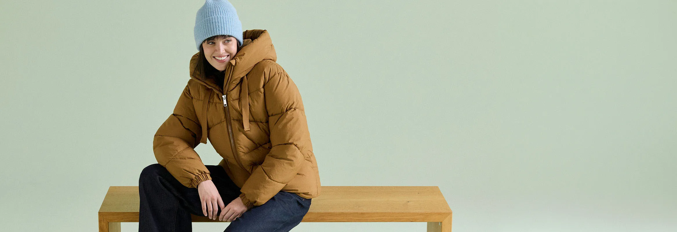 Une femme porte un bonnet bleu clair, une veste matelassée marron et un jean foncé.