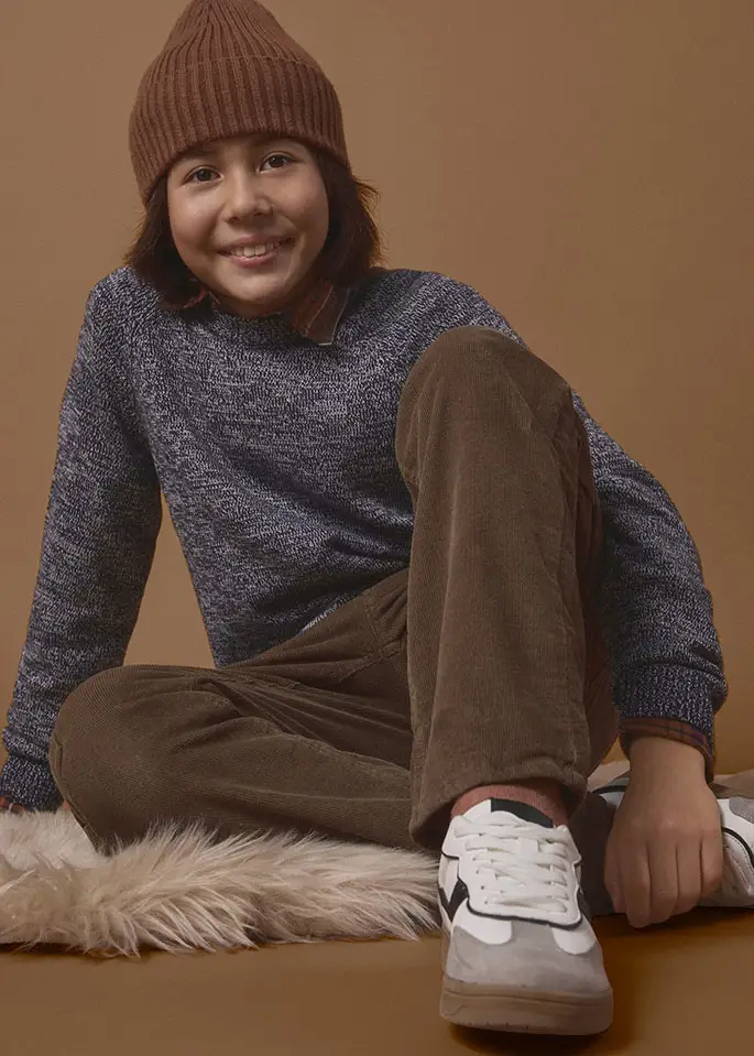 Enfant portant un bonnet marron, un pull chiné, un pantalon marron et des tennis blanches.