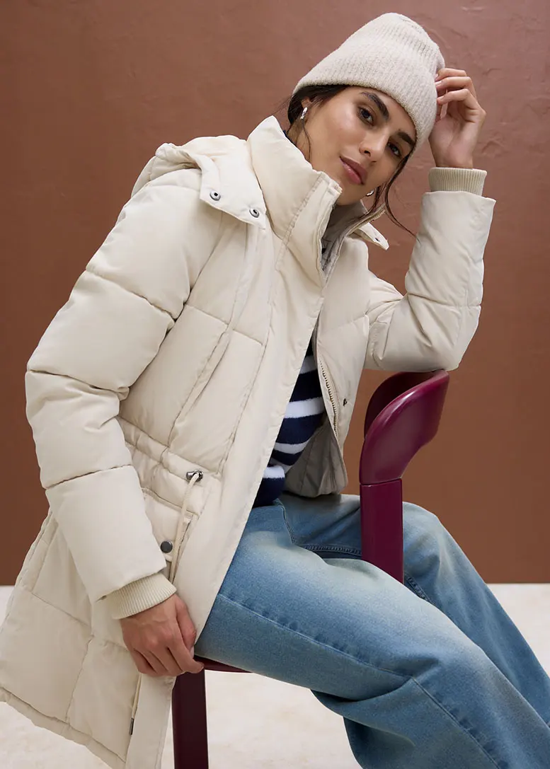 Une femme portant une doudoune beige, un T-shirt à rayures, un jean et un bonnet.