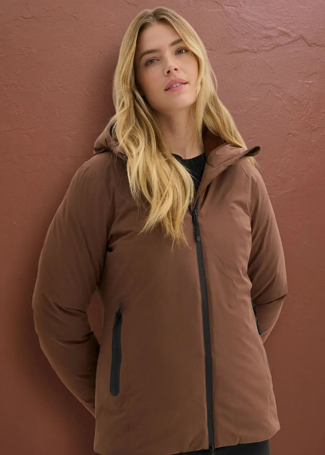 Femme portant une élégante veste matelassée marron à capuche et fermetures éclair noires.